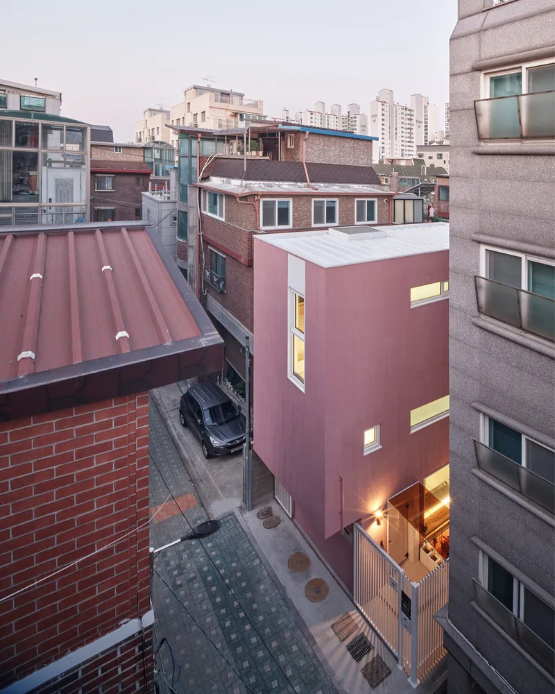 From above at dusk: pink house among grey and brown neighbourhood rooftops, windows glowing warm