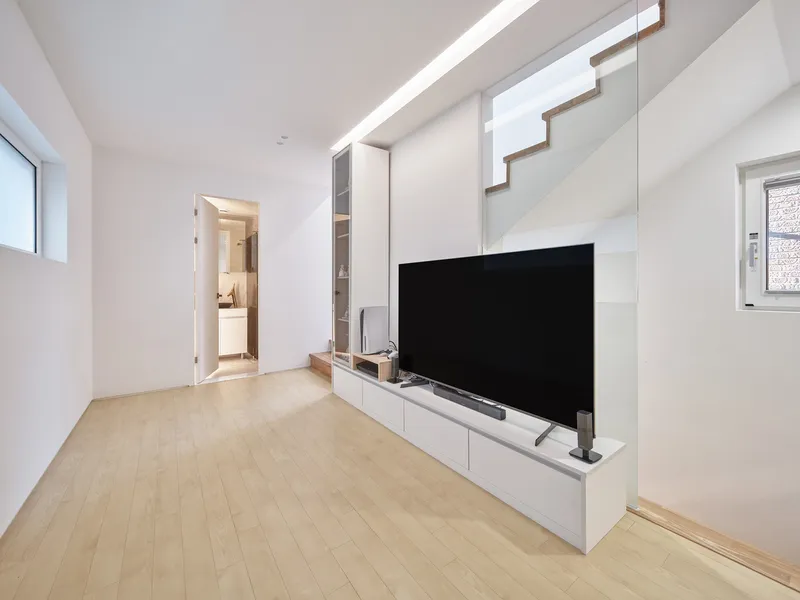 Living room: white walls, TV on low white cabinet, linear ceiling light, angular white stair balustrade, timber floor
