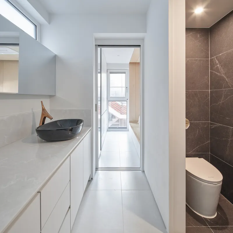 Bathroom: white counter with dark stone vessel basin, grey stone wall tiles, toilet, narrow window at the end