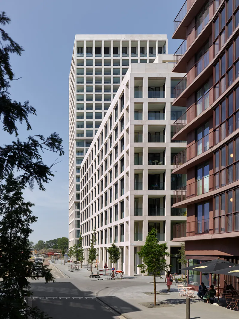 Street-level view of the white tower facade alongside brick residential building with young trees in foreground