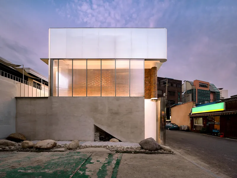 Street facade with concrete base, translucent upper volume and timber screens illuminated at dusk