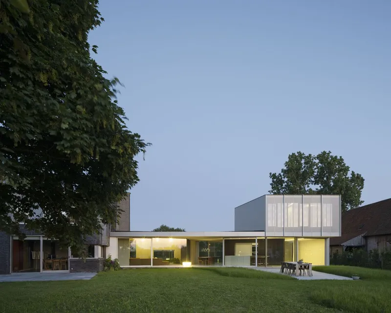 Rear elevation at dusk with illuminated glazed living spaces and perforated metal upper volume beside lawn