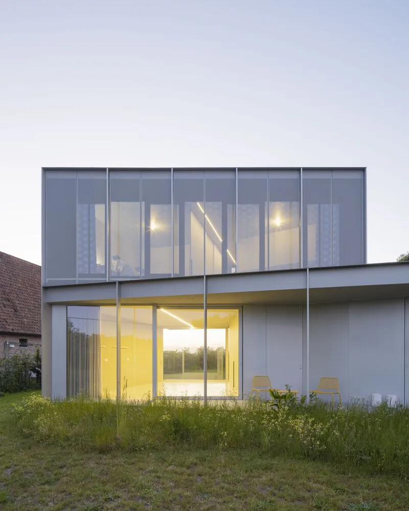 Two-story glass facade with perforated metal screens at dusk in an unmowed grassy field