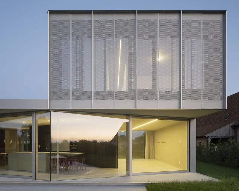 Upper level perforated metal panel facade above floor-to-ceiling glazing at twilight