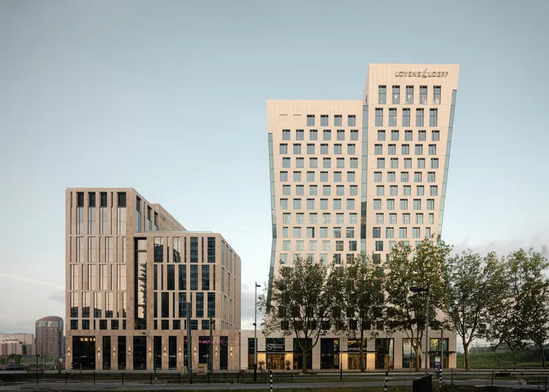 Streetscape view of two multi-story towers with beige precast panel facades and a row of mature trees