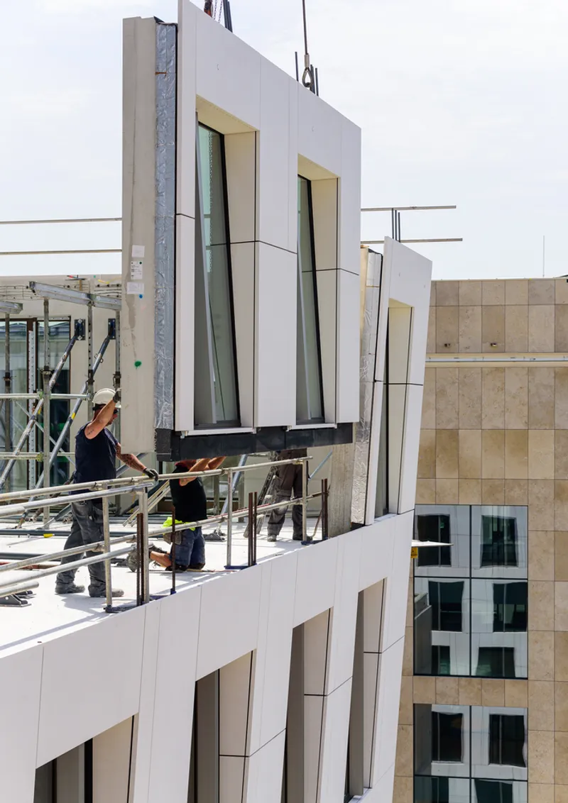Rooftop construction scene with workers installing white prefabricated facade panels with angled window recesses