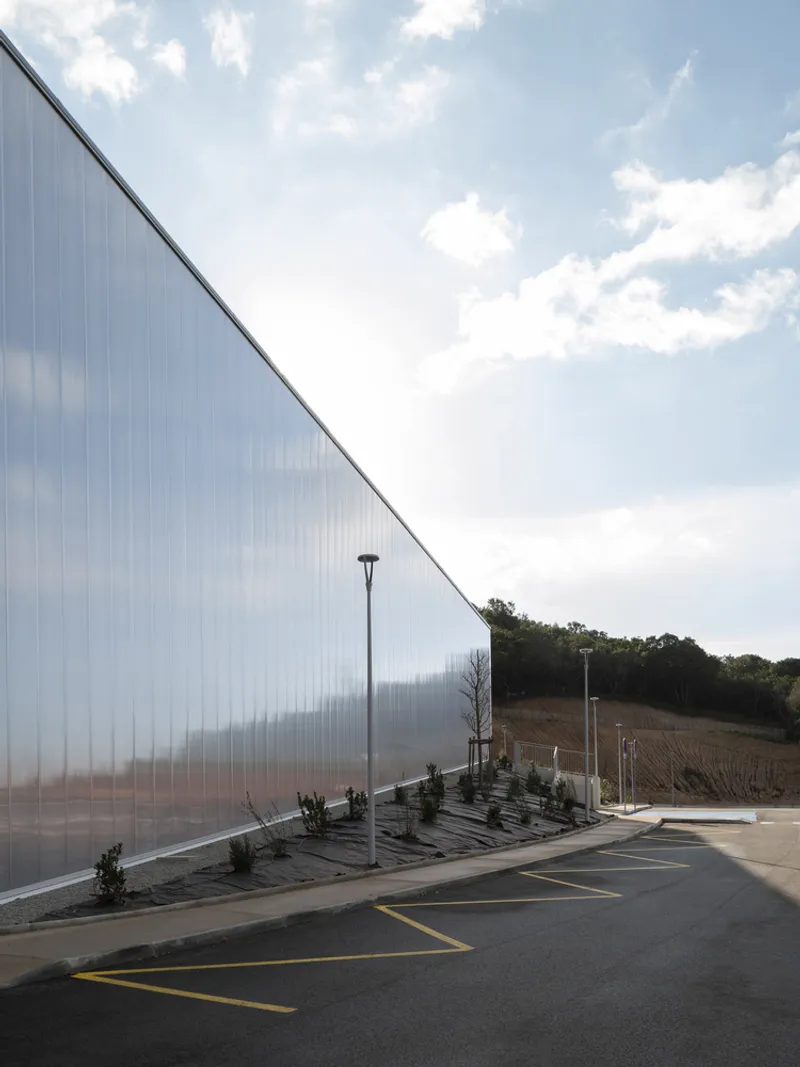 Angled view of the translucent polycarbonate facade with planted bed and parking lot under midday clouds