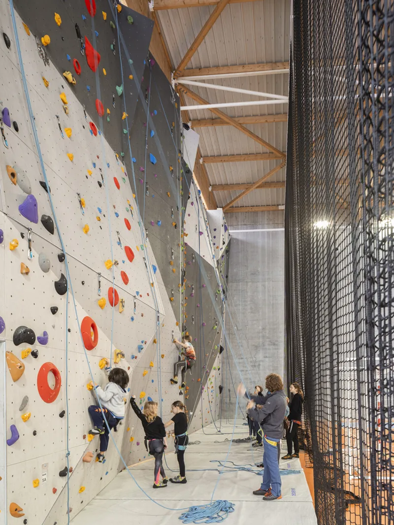 Indoor climbing wall with colorful holds and several climbers beside mesh safety netting under timber trusses
