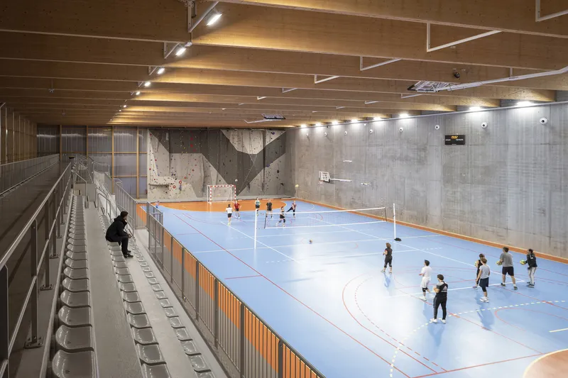 Gymnasium with bleacher seating, board-formed concrete walls and children playing on the blue sport court