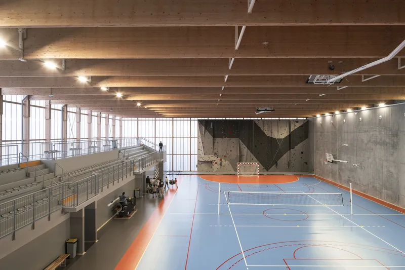 Multi-court sports hall with exposed timber beams, bleacher seating, and climbing wall with geometric pattern