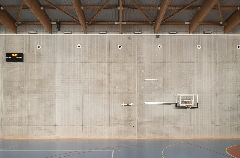 Board-formed concrete wall with basketball hoop and timber roof trusses above a blue court