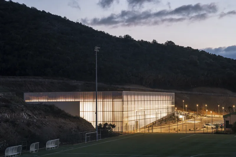 Translucent panel facade glowing at dusk beside a sports field with forested hillside beyond
