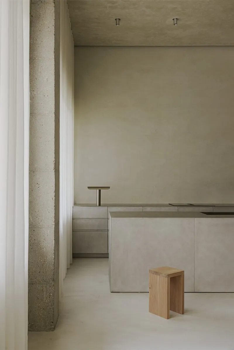 View through concrete columns toward a pale plastered kitchen counter with a timber stool on polished flooring