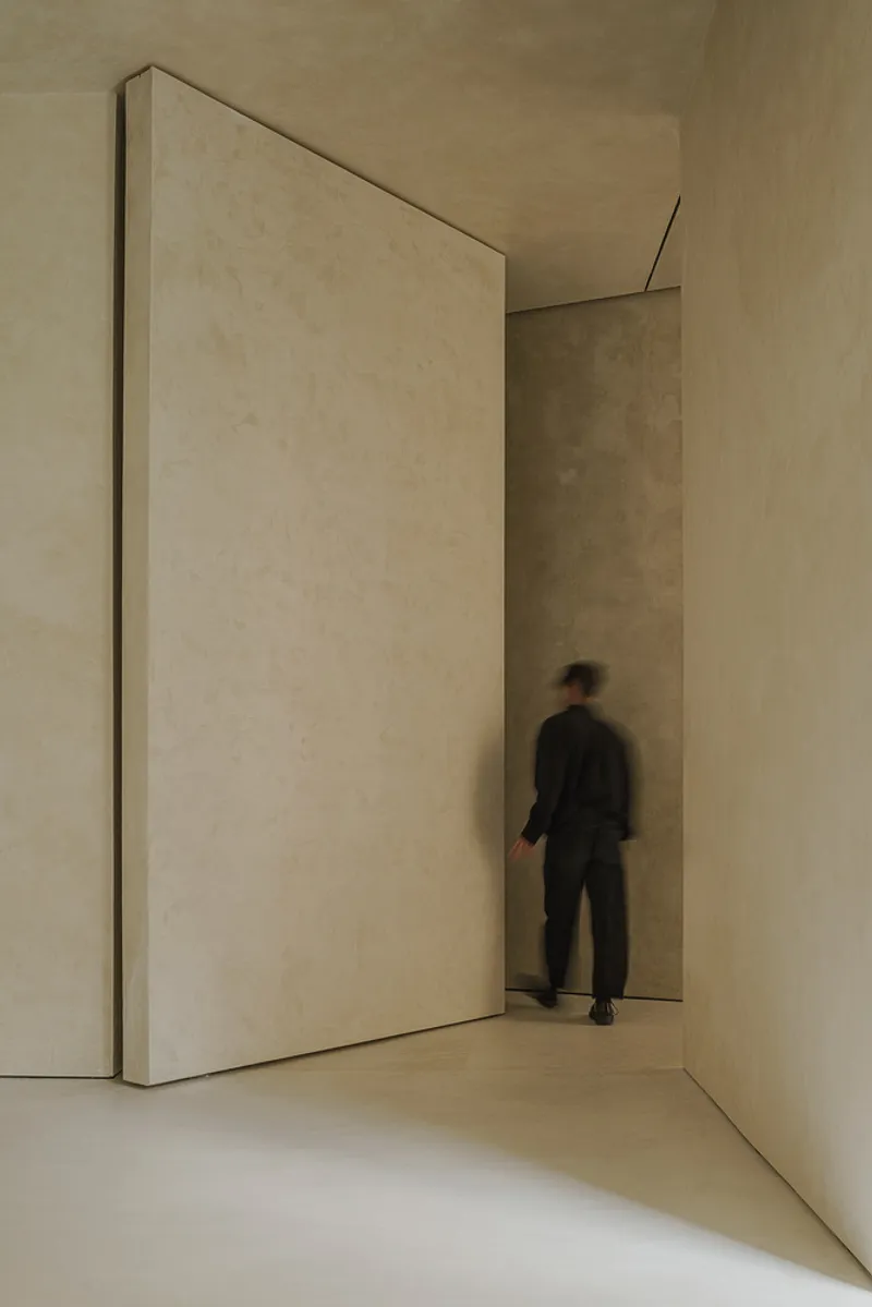 Tall pivoting plaster-clad door with a person walking through the narrow passage in soft light