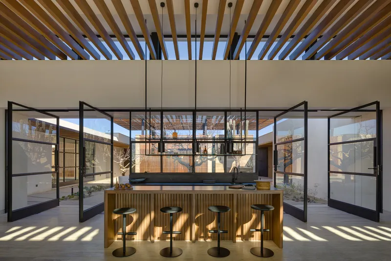 Kitchen with courtyard doors: timber island with black pendant lights, full-height black-framed pivot glass doors opening to the courtyards on both sides, slatted timber ceiling