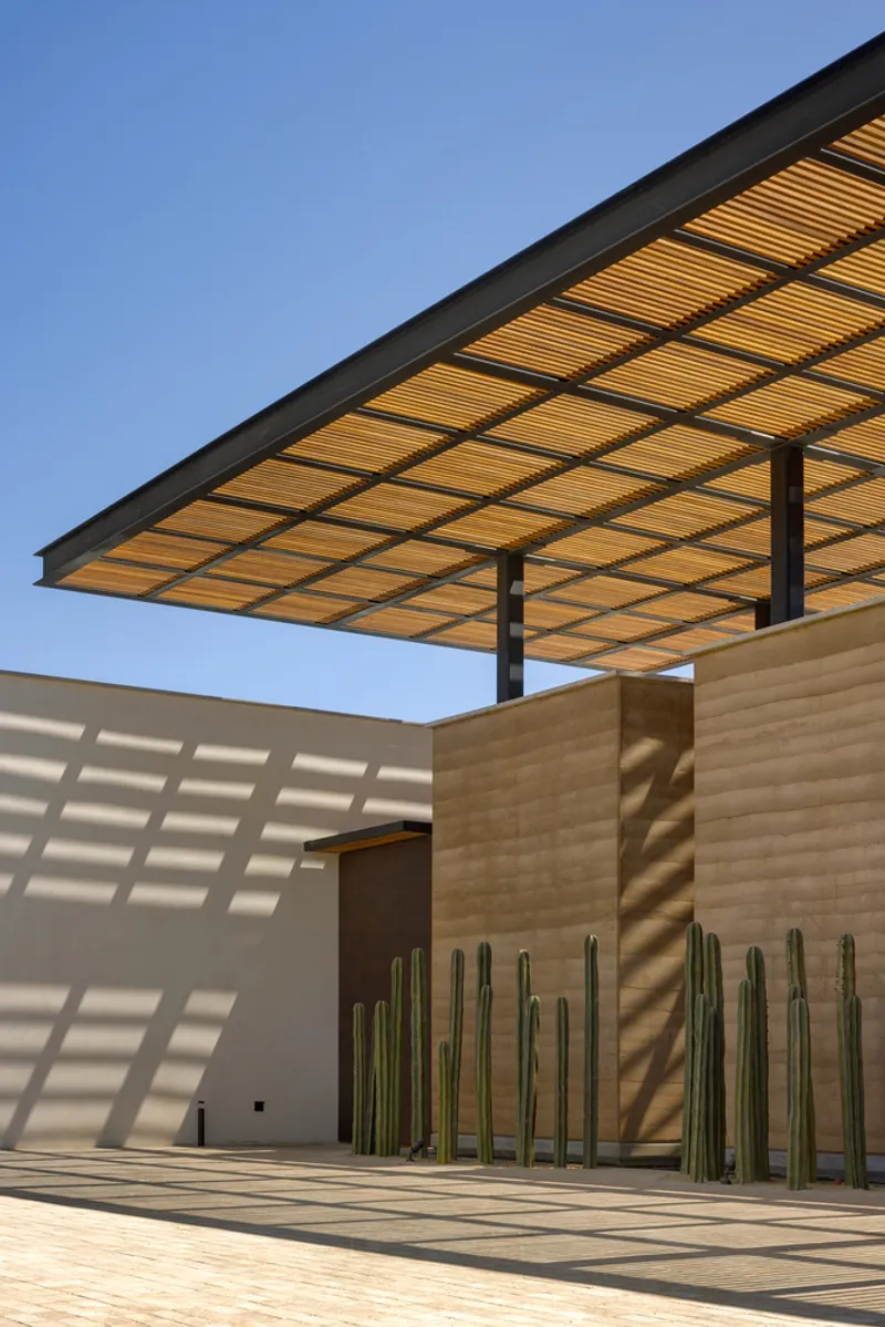 Facade detail: rammed earth wall with a timber pergola casting diagonal shadows, vertical timber slat screen, cactus silhouette on the wall