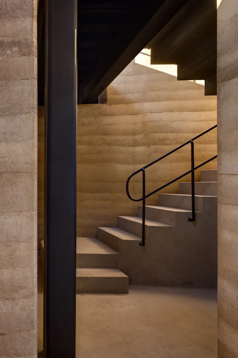 Stair: rammed earth walls with visible layered strata, concrete steps with a slender black steel handrail, view through to an adjacent room