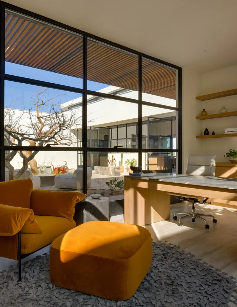 Study: orange velvet armchair with ottoman, black-framed window wall to the desert, timber desk, pivot door to the courtyard, exposed timber ceiling