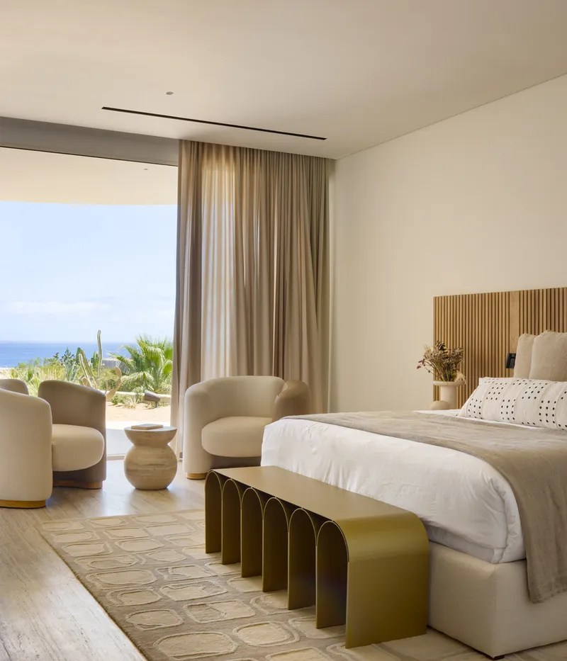 Master bedroom: upholstered headboard, pale linen bedding, curtained glass wall opening to a terrace with a view of the sea and distant headlands