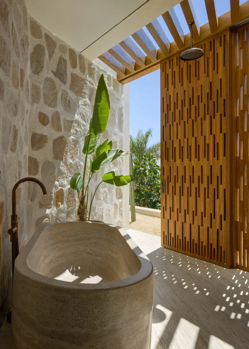 Outdoor shower: rough stone wall, carved stone soaking tub, timber slat door, banana plant, overhead timber pergola letting light through