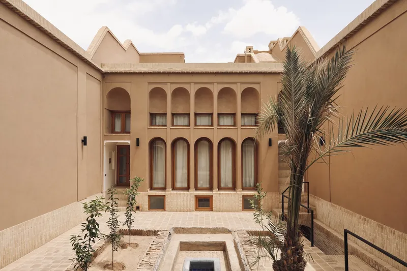 Courtyard with arched facade, sunken pool, palm trees and brick paving under partly cloudy skies