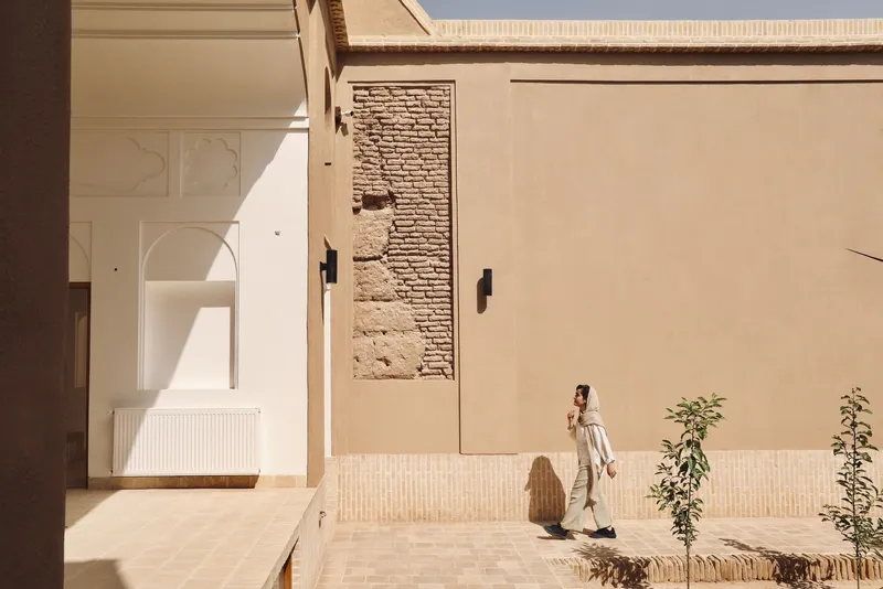 Courtyard passage with figure in white walking past exposed brick and tiled base in afternoon light