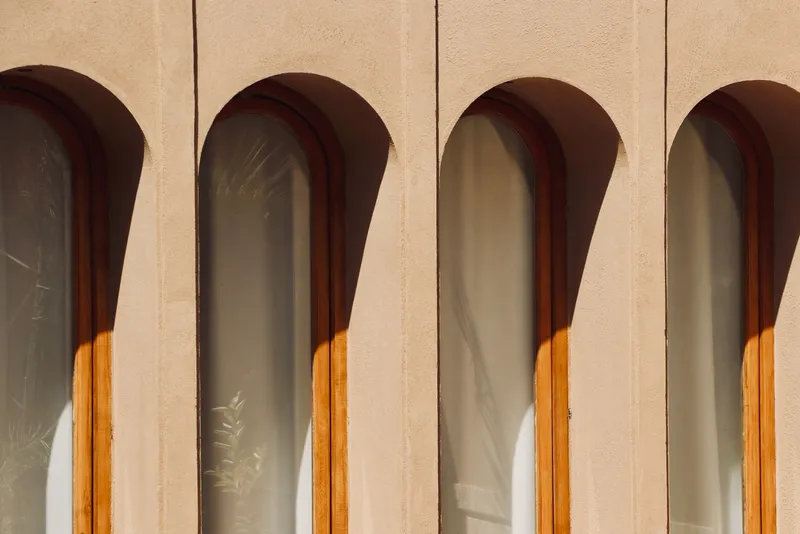 Row of arched windows with timber frames set into a plastered facade in direct sunlight