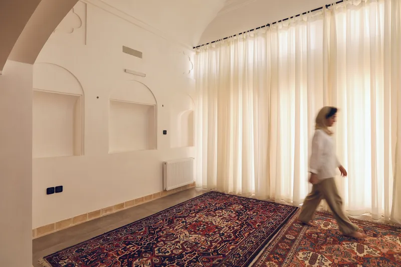 White room with arched wall niches and a figure walking past sheer curtains in afternoon light