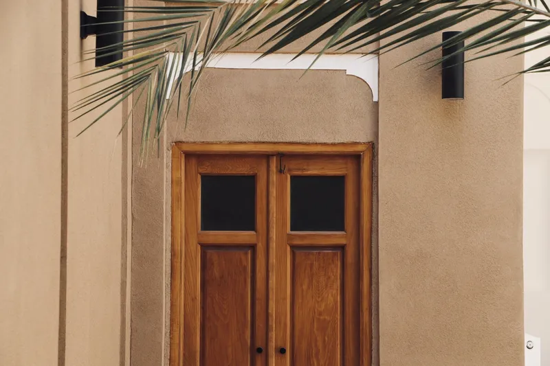 Timber double door with arched opening beneath a palm frond in a stucco wall