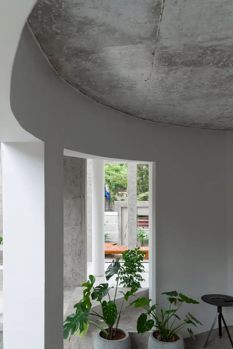 Interior view through doorway to concrete columns and outdoor courtyard with potted monstera plants below curved soffit