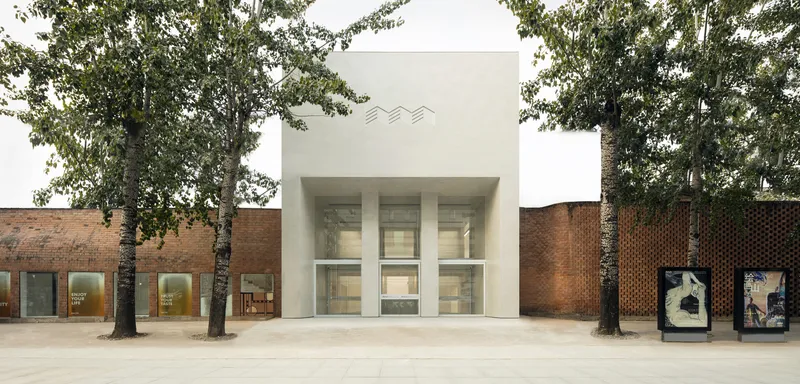 Street facade: white cubic volume rising above red brick neighbours, wavy logo, three tall glass openings, plane trees framing the entry