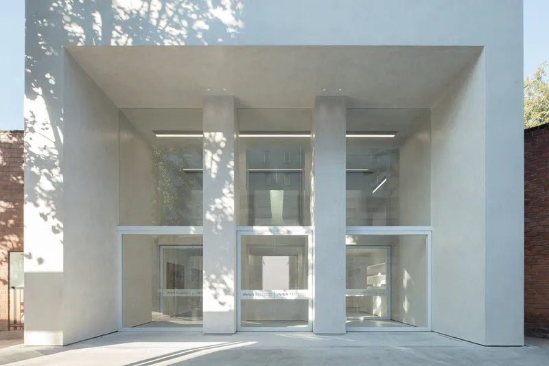 Entry close-up: three full-height glass panels between white concrete columns, tree shadows on the facade, double-height lobby visible