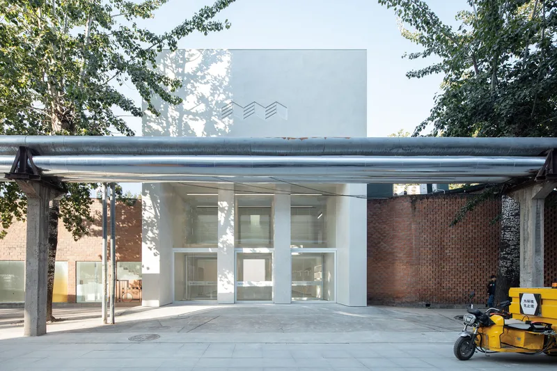 Street context: white entry volume with wavy logo, metal canopy, red brick neighbours, plane trees, delivery scooter at right