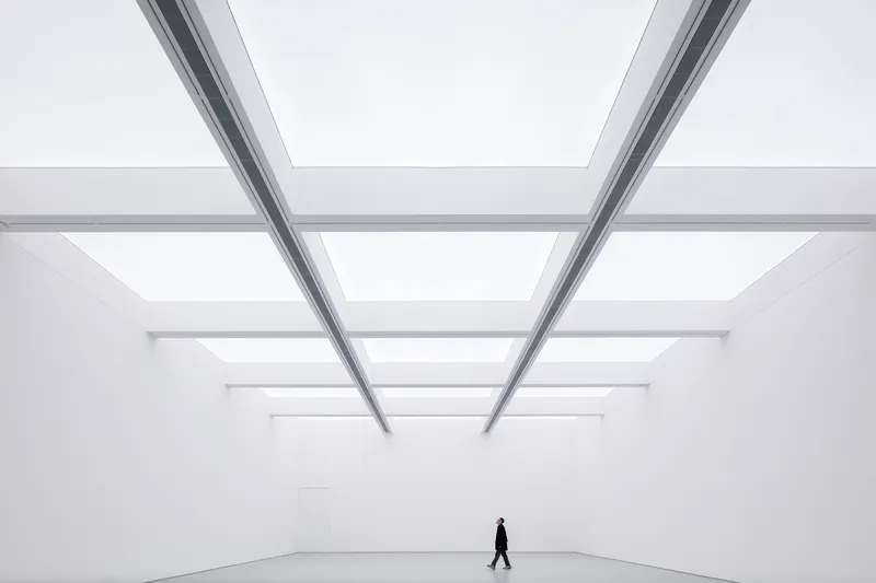 Main hall with figure: person walking across the vast white room under a skylight grid, steel beams converging above