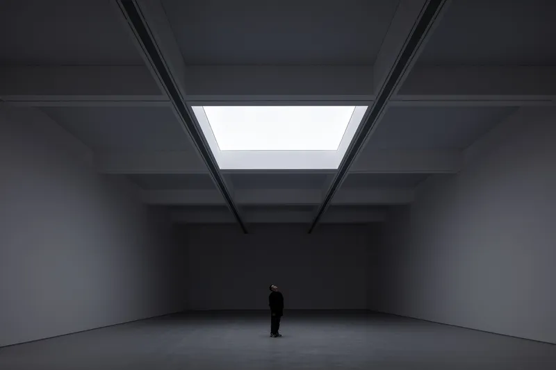 Darkened hall: single central skylight illuminating a dark exhibition space, figure standing below