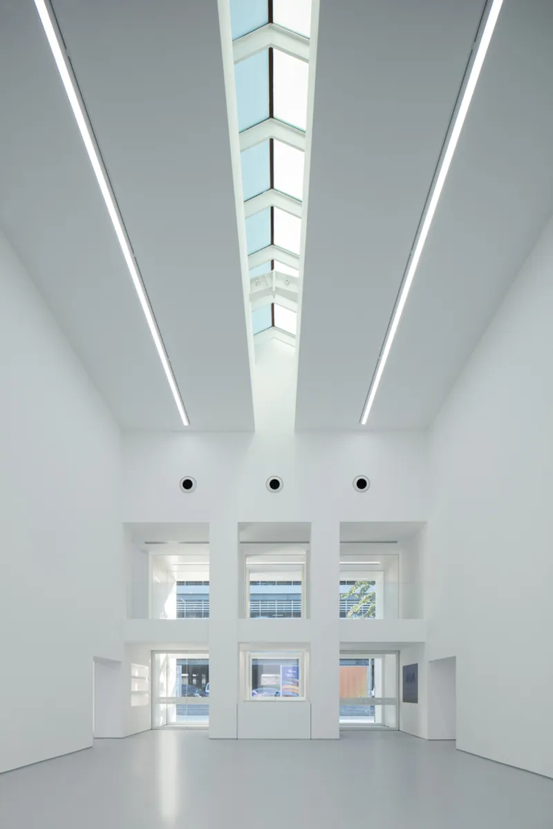 Lobby interior: tall white volume, skylight strip running the length of the ceiling, three windows to the street at the far end