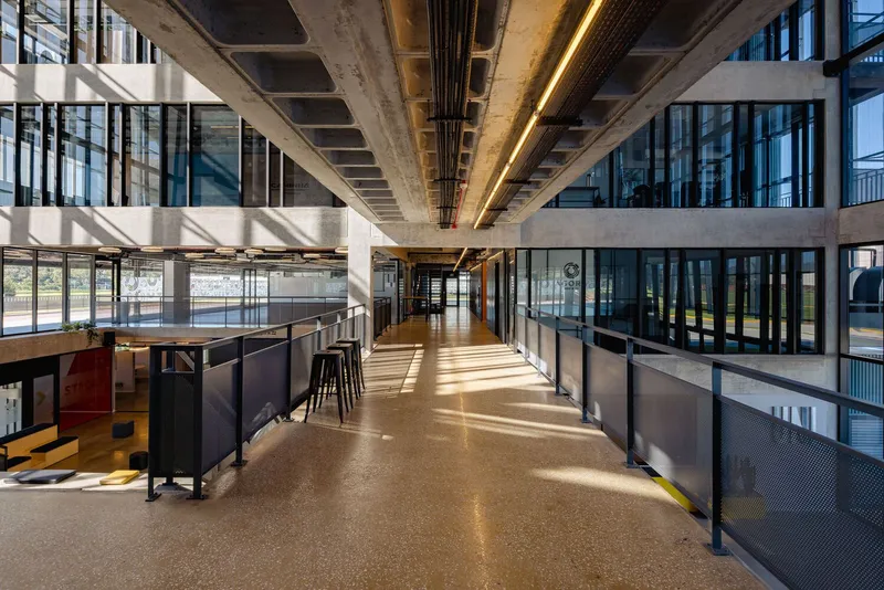 Interior corridor with exposed concrete beams, timber ceiling panels, and sunlight filtering through glazed walls