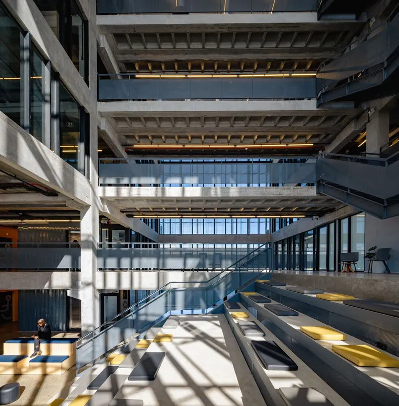 Multi-story void with stacked concrete floors, steel stairs and sunlight streaming through glazed facade
