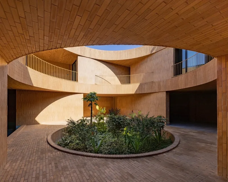 Central courtyard photographed from ground level: a circular void carved through the house reveals a raised planter at the base, warm brick-clad walls, and a curved upper balcony overlooking the greenery
