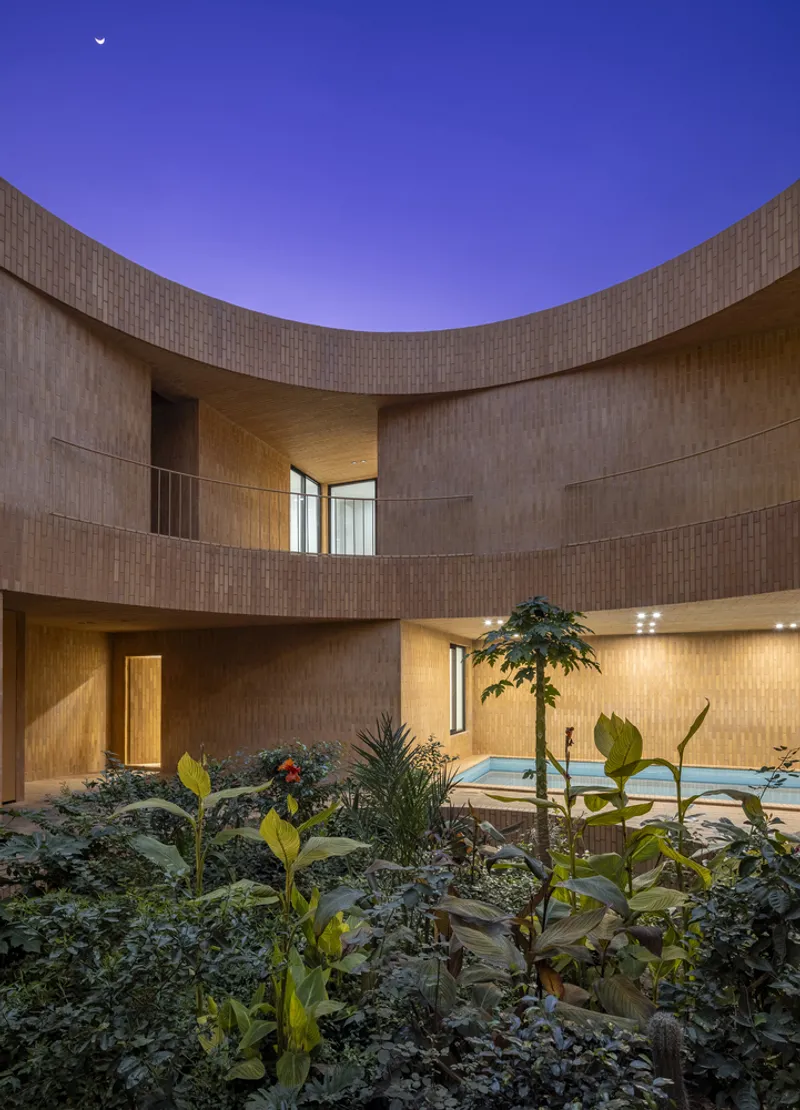 Courtyard at dusk: the curved brick wall wraps around lush planting at the base, with interior rooms glowing warmly against a deep purple evening sky