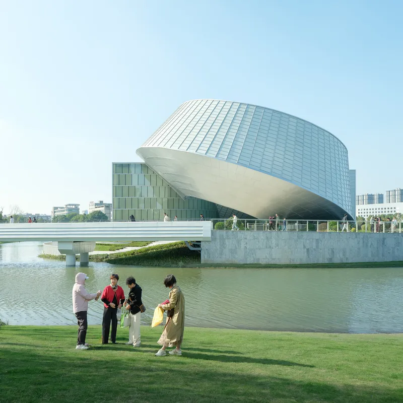 White dome with gridded glazing and tilted block facade beside a concrete bridge with visitors on lawn