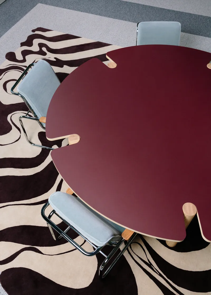 Top-down view of maroon table surface with blue chairs on carpet with swirling graphic pattern