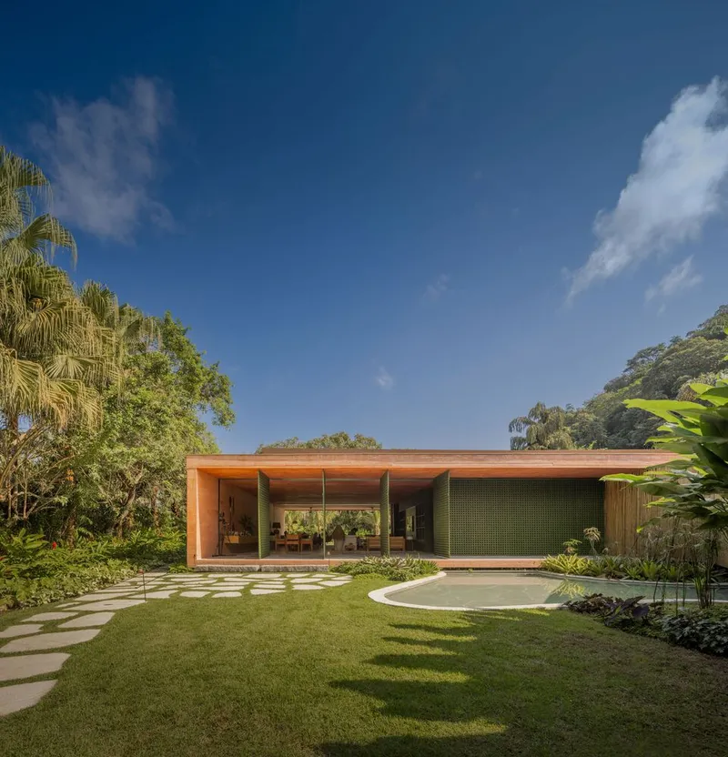 Single-story timber pavilion with large glazing opening onto a lawn and curved paver path under blue sky