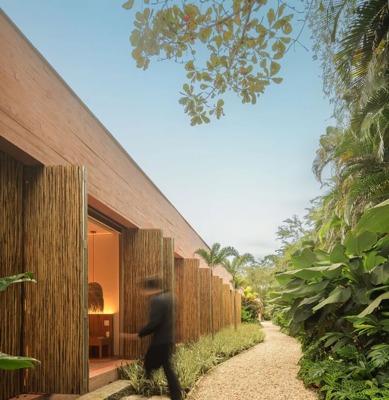 Bamboo-clad facade with illuminated doorway and figure walking along gravel path at dusk