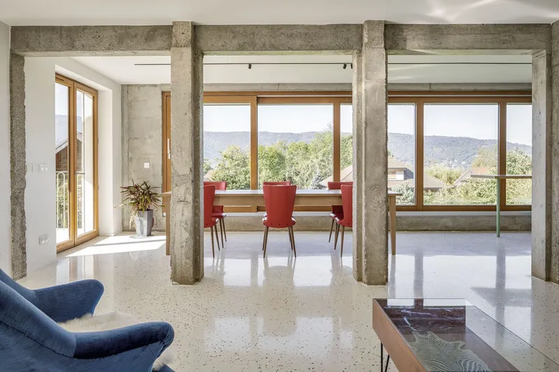 Through the columns: concrete structure framing the dining area with red chairs, full-height timber windows, mountain view