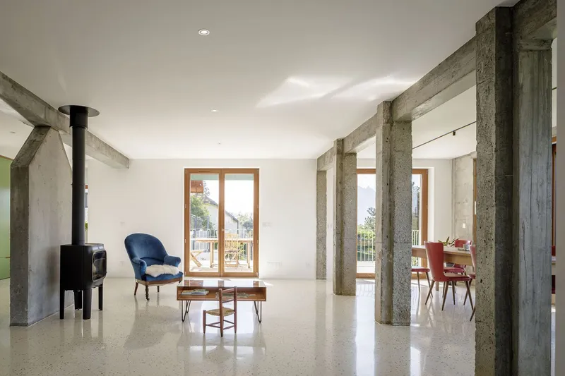 Open-plan living: exposed concrete columns and beam, terrazzo floor, wood stove, blue velvet chair, dining beyond