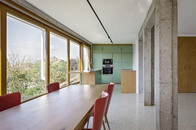 Kitchen and dining: sage green cabinets, long timber table with red chairs, concrete column, mountain view through windows