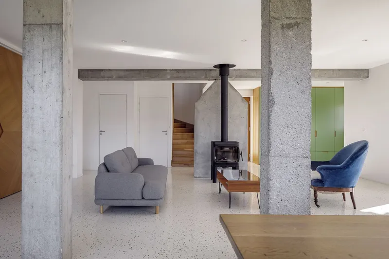 Living room: exposed concrete columns and beam, grey sofa, blue chair, wood stove, green kitchen beyond