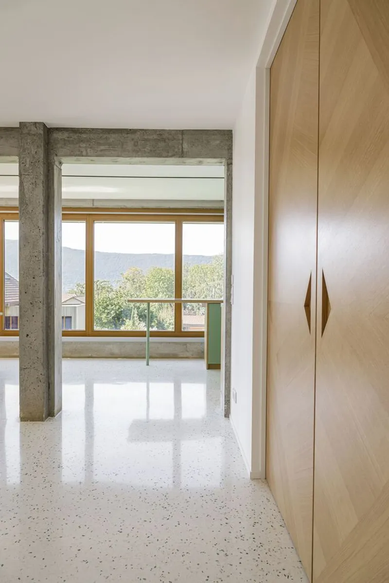 Corridor: timber wardrobe, concrete beam overhead, terrazzo floor, green kitchen glimpsed through the opening