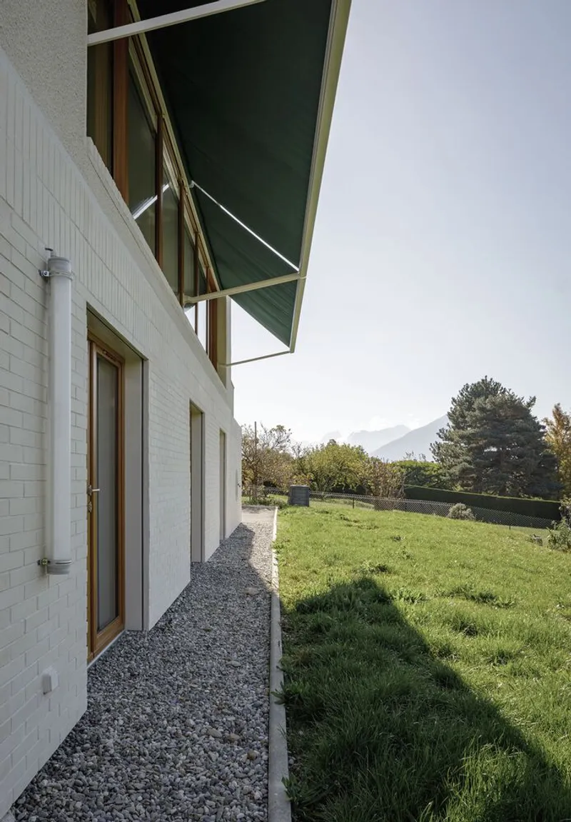 Side passage: green awning overhead, gravel path along white rendered wall, garden beyond